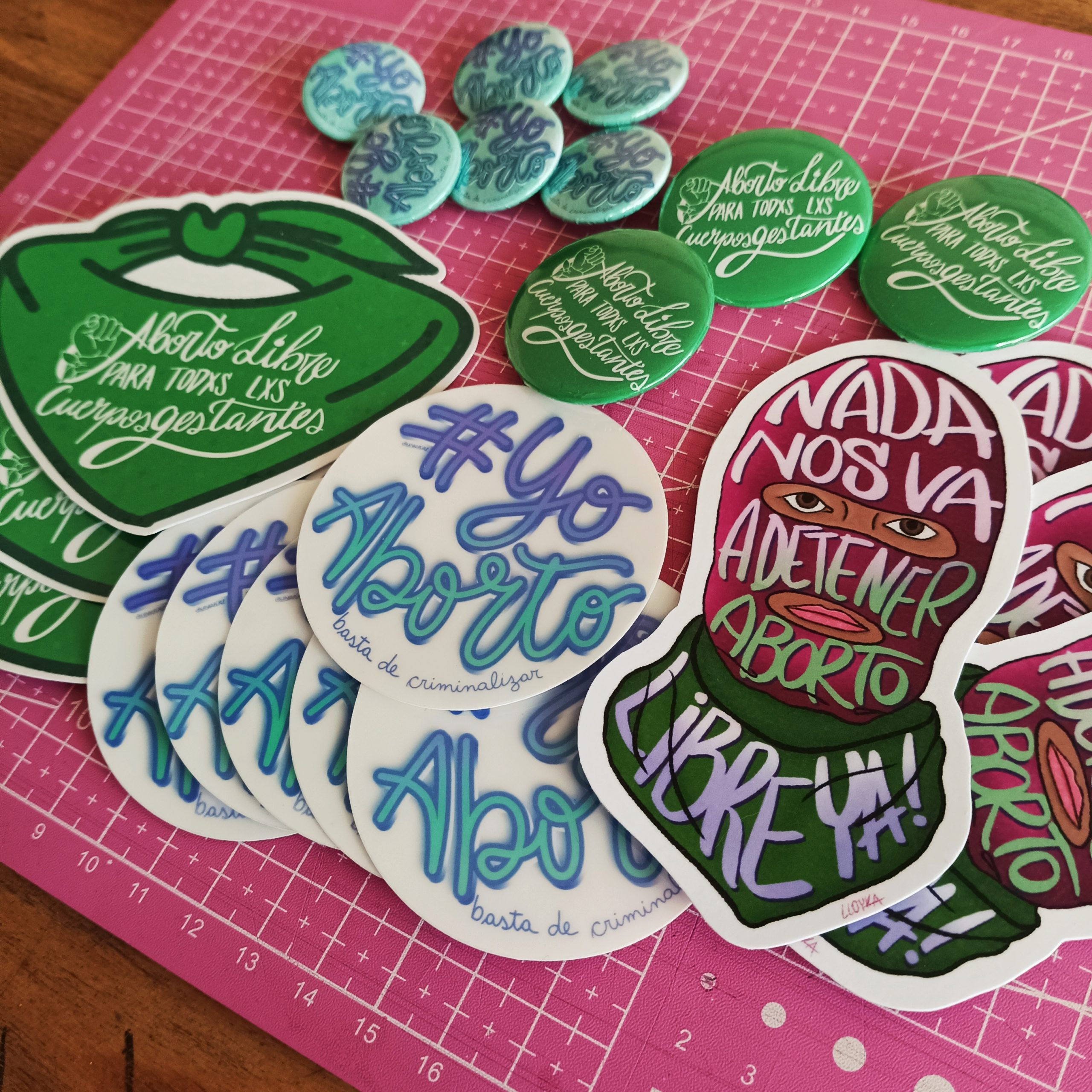 Una colección de pegatinas y pins a favor del derecho al aborto se extienden sobre una alfombra de corte rosa. Los diseños presentan letras e ilustraciones llamativas en tonos verdes, azules, morados y rojos.
