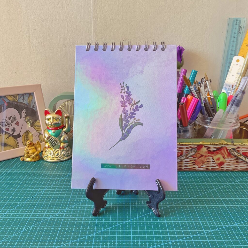Libreta anillada vertical con ilustración de tapa trasera de lavanda sobre fondo acuarela en tonos lilas, celestes y rosados. Está sobre un atril negro, con un escritorio de fondo decorado con lápices, reglas, figuras de la suerte y una ilustración enmarcada.