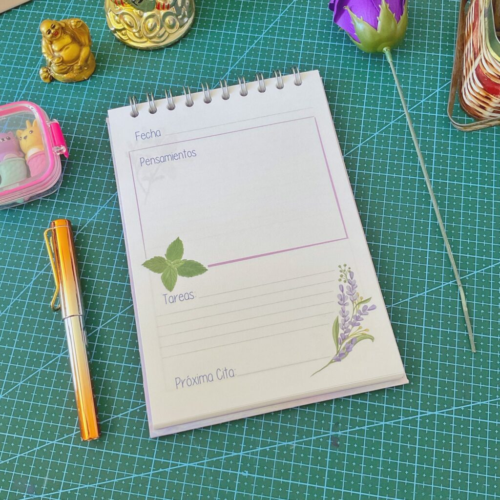 Página interior del cuaderno de terapias con secciones tituladas “Fecha”, “Pensamientos”, “Tareas” y “Próxima Cita”, decorada con ilustraciones de lavanda y hojas verdes. Está sobre una base de corte verde con objetos como un lapiz naranja y figuras decorativas alrededor.