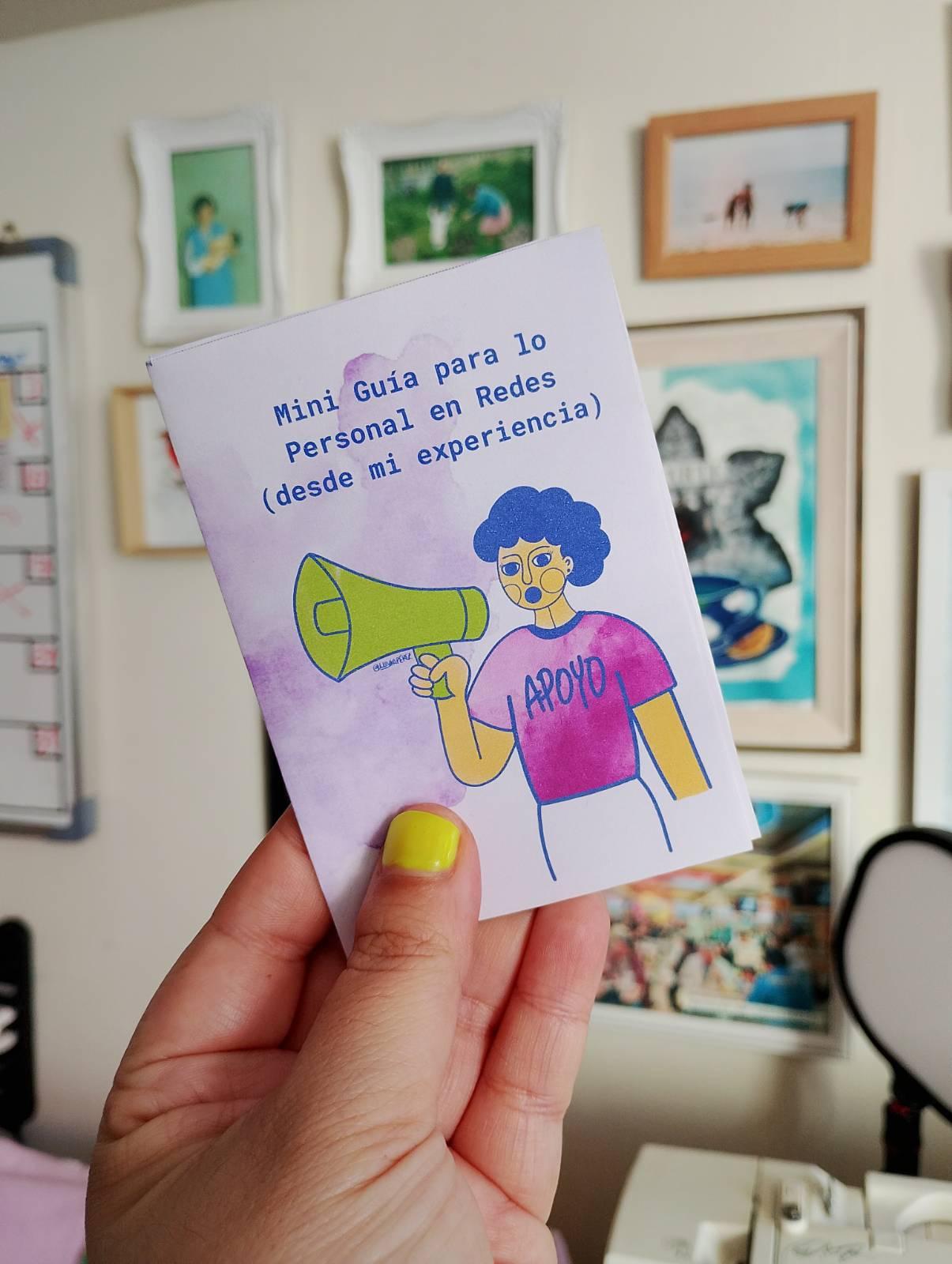 Una mano sostiene un pequeño fanzine titulado “Mini Guía para lo Personal en Redes (desde mi experiencia)”. La portada está ilustrada con una persona de cabello azul que sostiene un megáfono verde. Lleva puesta una polera morada con la palabra “APOYO”.