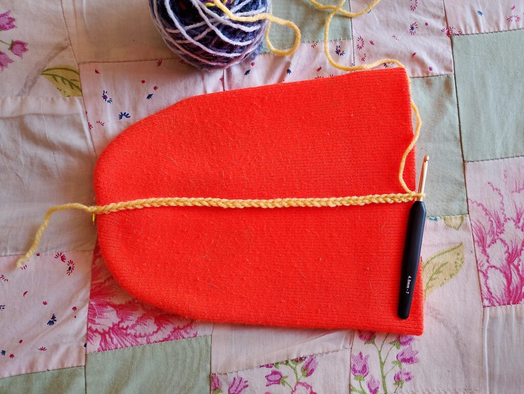 Fotografia donde se encuentra un gorro de lana tipo benie naranjo con el cual sacaremos las medidas de inicio para tejer nuestro gorro, sobre el se encuentra la cadena de puntos de inicio en lana amarilla y una aguja de ganchillo encima. Cerca hay un ovillo de lana morada y blanca, todo ello sobre una colcha.