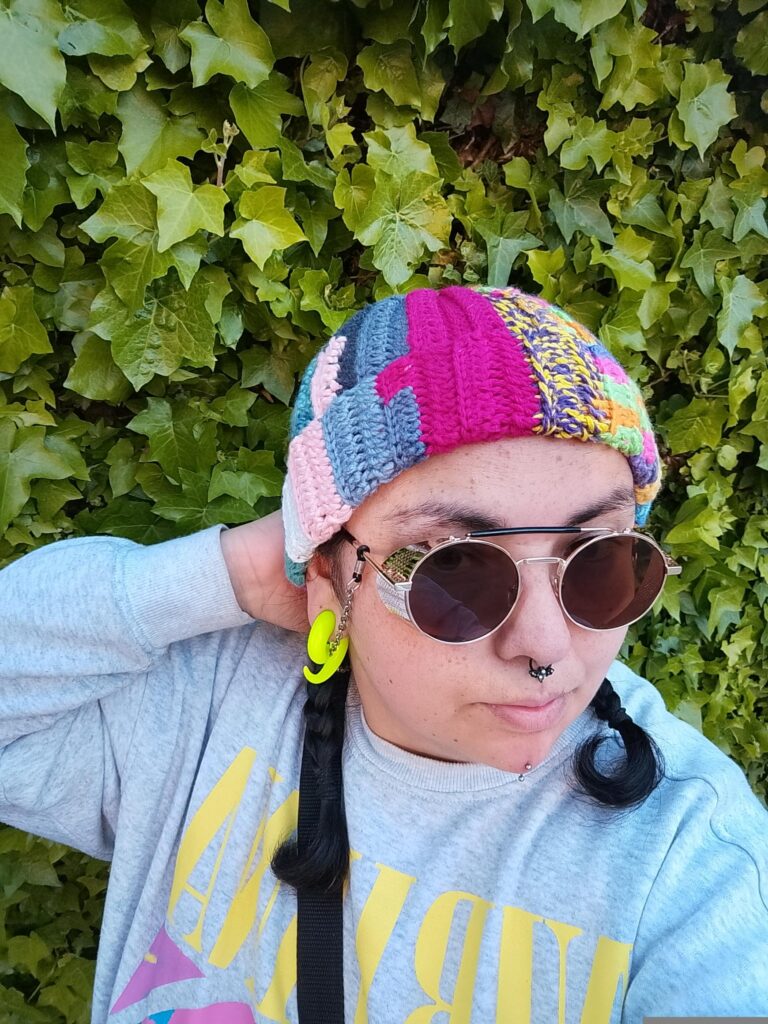 Una persona con trenzas, que lleva gafas de sol redondas, tiene puesto el gorro de punto de colores y una sudadera clara posa delante de una hiedra verde. Lleva pendientes amarillos brillantes.