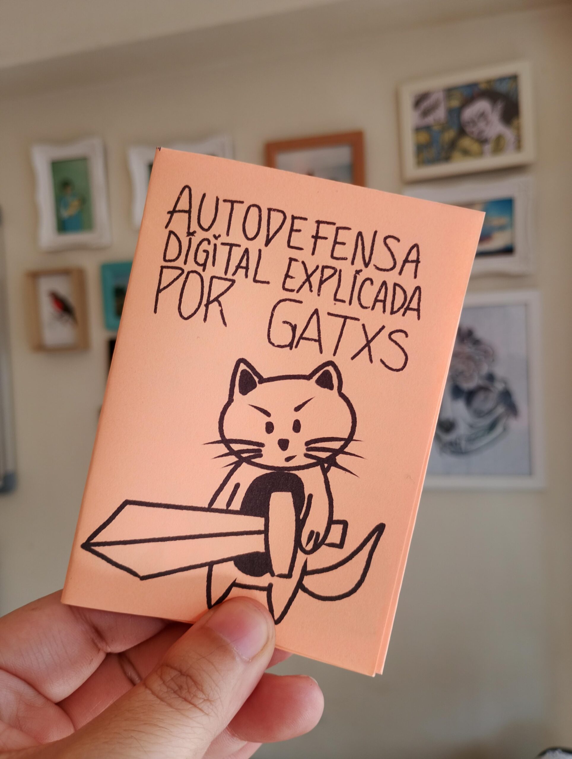 Mano sosteniendo un pequeño fanzine color durazno que dice ‘Autodefensa digital explicada por gatxs’. En la portada hay un dibujo de un gato chibi sujetando una espada. Al fondo, se ven cuadros colgados en una pared desenfocada.