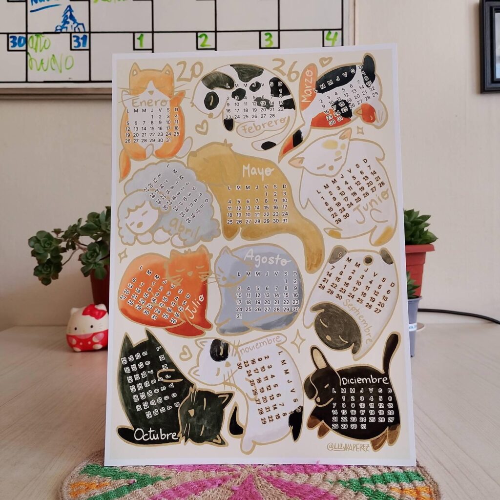 Calendario 2026 impreso en papel, ilustrado con múltiples gatos de distintos colores y formas. Cada gato contiene un mes del año dentro de su silueta. El calendario está apoyado sobre una mesa clara con una planta suculenta al fondo.