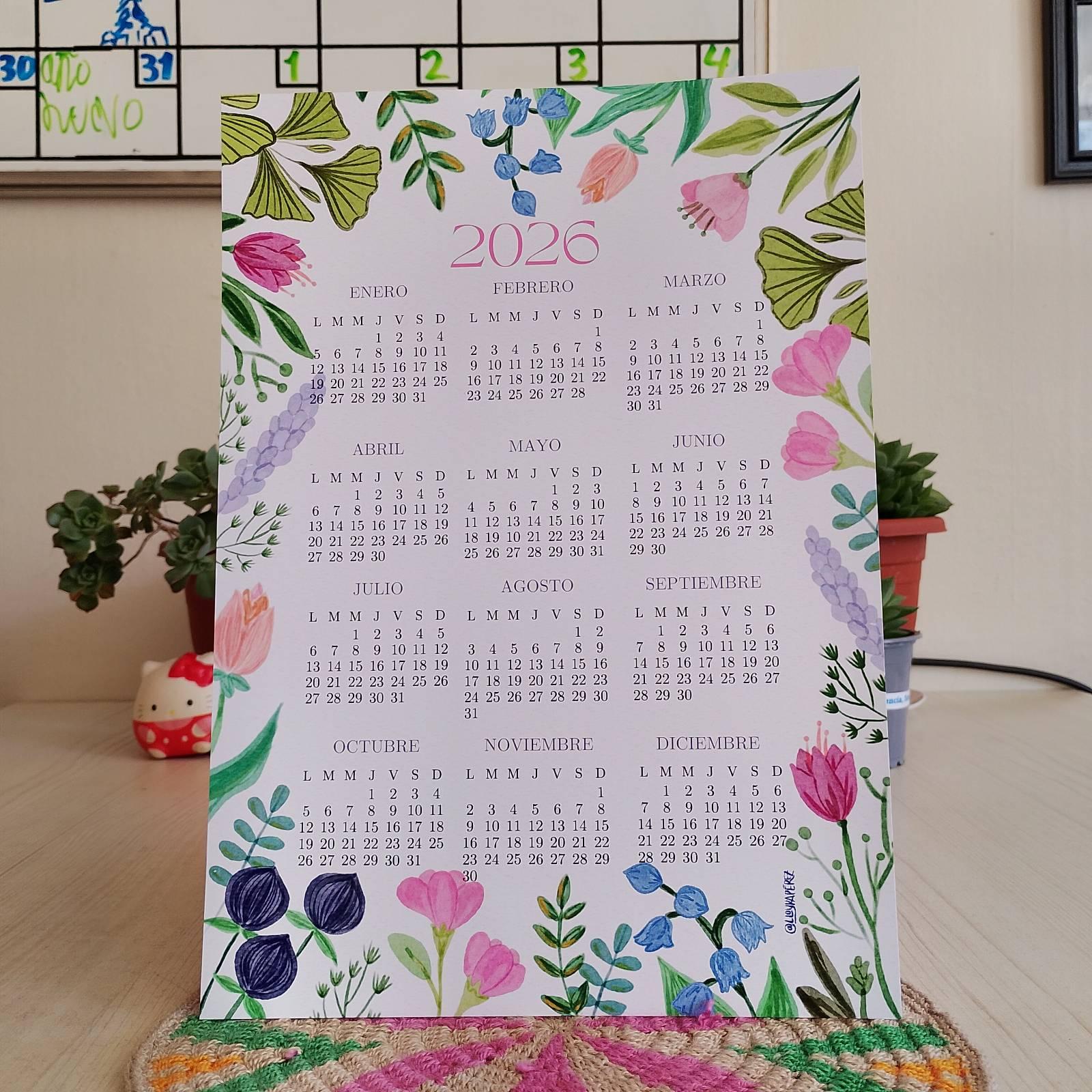 Calendario 2026 impreso en papel con un diseño floral en acuarela alrededor del borde. En el centro están los meses del año. El calendario está apoyado sobre una mesa clara, con plantas en macetas al fondo.