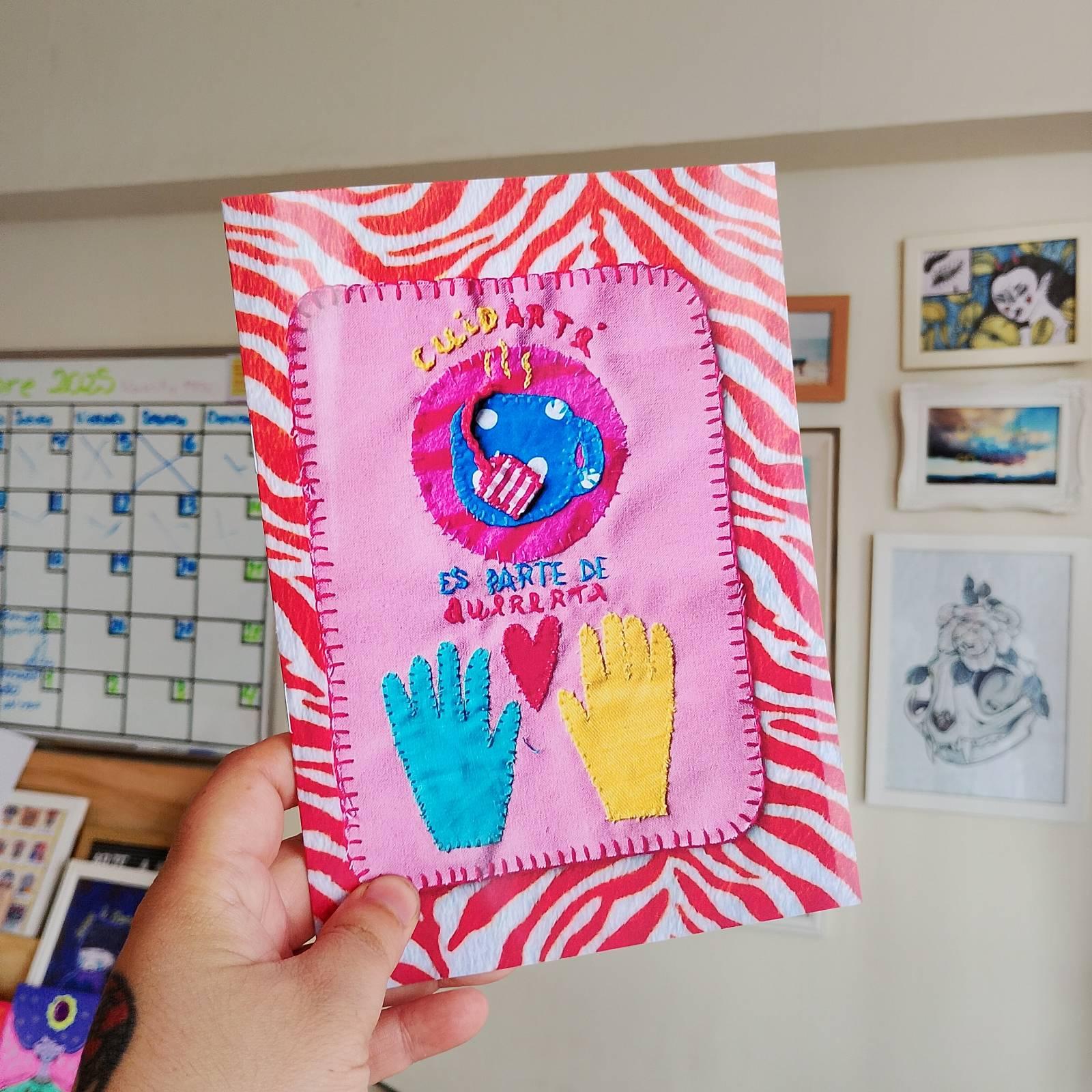 Portada del fanzine Cuidar Te sostenido con una mano. Fondo con patrón de rayas rojas y blancas tipo cebra. Al centro hay un parche textil bordado en tonos rosados con la frase “CuidarTé es parte de Quererte”. Incluye un bordado circular con una taza de te, dos manos de colores azul y amarillo, y un corazón rojo entre ellas. De fondo se observa una pared con ilustraciones enmarcadas.